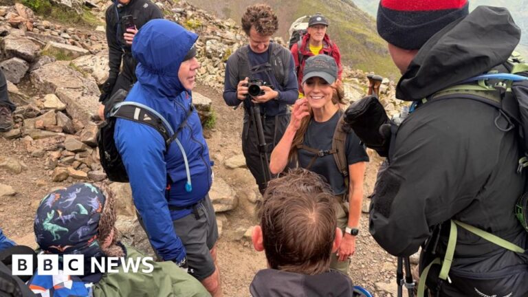 A woman in smiling on a mountain surrounded by fellow walkers all wearing winter and waterfproof clothing.