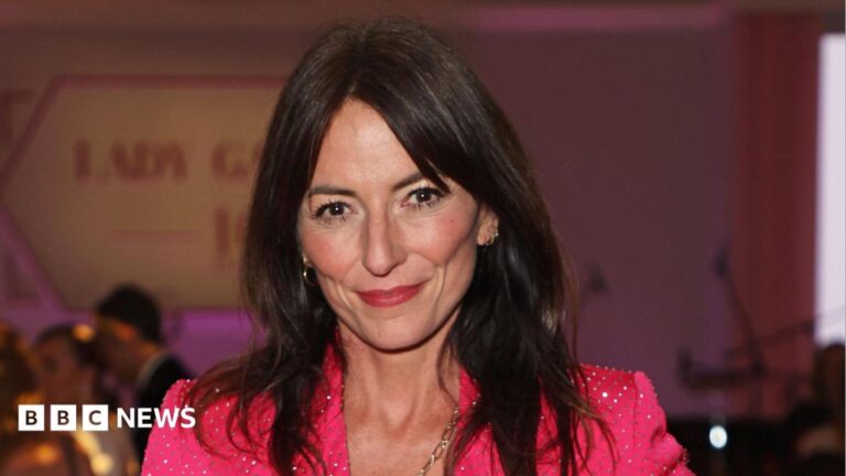 Davina McCall in 2024 is smiling at the camera. She has shoulder-length dark hair and is wearing a sparkly pink jacket.