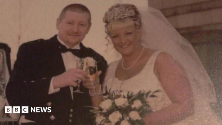 A man in traditional Scottish dress including a black bow-tie, waistcoat and kilt, stands next to a woman in a white wedding dress and veil. Both are smiling and are clinking glasses of champagne.