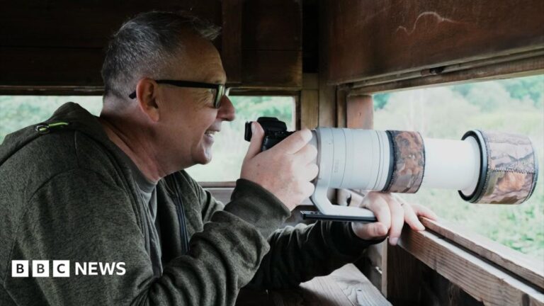 Alan Bruce is sitting in a wooden wildlife hide. He has a long-lens camera resting in front of him, sticking out of the open side of the hide. Alan is wearing a green hoodie and has dark-rimmed glasses