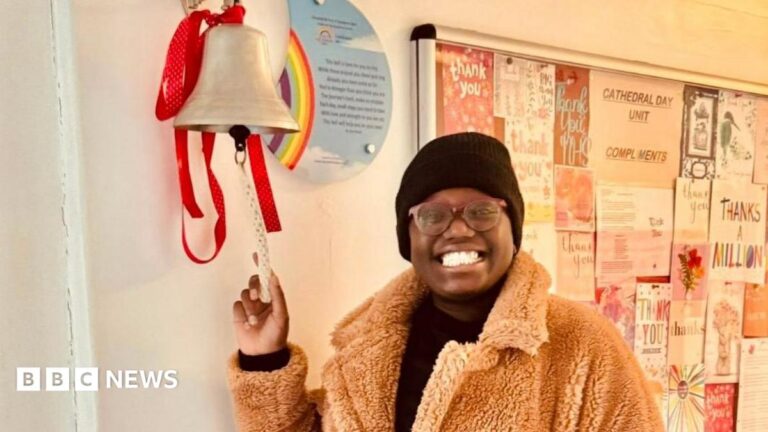 A woman smiling at the camera. She is wearing a black hat and brown fluffy coat. She is ringing the 'remission' bell in the hospital.