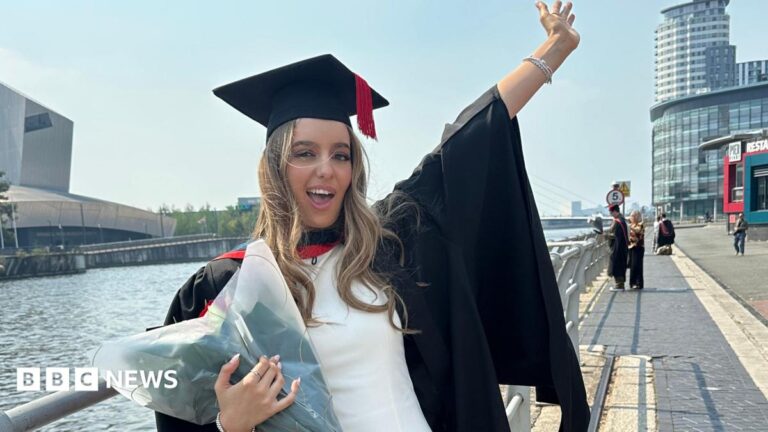 ‘I didn’t think I’d be graduating’ after Freshers’ Week cancer op Ella is standing in her graduation gowns next to Salford Quays. She is holding flowers in one hand and waving her other in the air. She has a joyful smile.