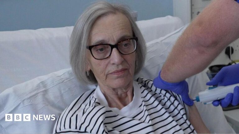Chris Jones receives a vaccine into her left arm. She is lying in a hospital bed and wears a white and black striped top. She has grey hair and wears dark-framed glasses.
