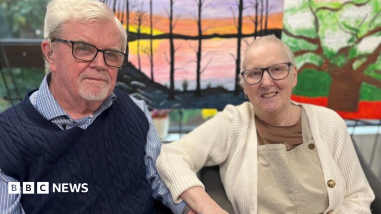 The special friendship formed while receiving palliative care Sally and Kevin are looking at each other and smiling. Kevin is wearing a blue and white striped shirt and a navy knitted tank top. Sally is wearing a cream cardigan, brown t-shirt and light brown dungarees. Both have short white hair and glasses.