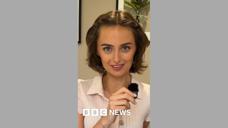 Young woman with short brown hair, smiles at camera, while holding a small clip mic in her right hand. She is wearing a pink short sleeved t-shirt.