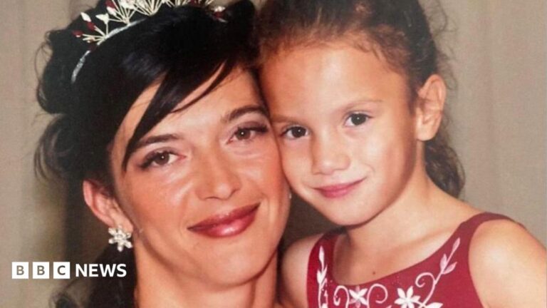 A woman with dark hair tied up with a silver tiara looks at the camera and cuddles a young girl with tied back dark hair and a red dress