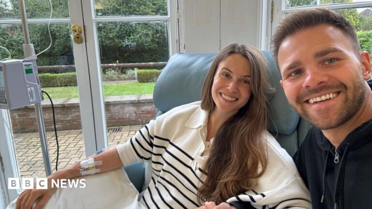 Claudia Adolphus-Green sits in a blue treatment chair with a cannula attached to one arm. Her husband Rob Green is sitting next to her and both are facing towards the camera. Large glass doors behind them look out onto a patio, lawn and landscaped garden.