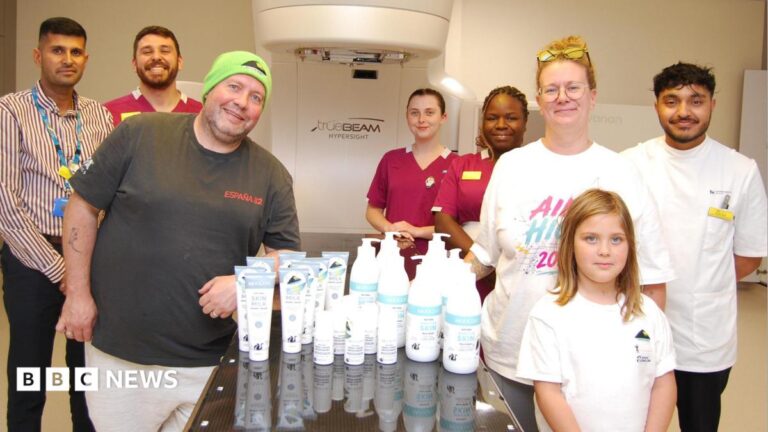 Charity donates skincare to radiotherapy patients in Northampton A dark glass table is at the centre of the picture. On top of the table are white and blue pots, tubs and bottles of skin care products. Seven adults and one child are standing around the table looking at the camera. Some are wearing hospital uniform, three are wearing white t-shirts. They appear to be in a radiotherapy room with a large piece of medical equipment behind them.