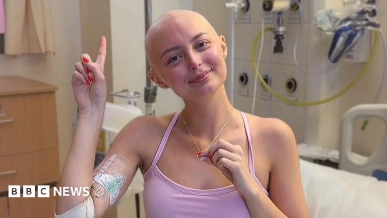 Erin survived cancer aged 21 – now it’s a race against time for a chance of a baby Person sitting in a hospital room wearing a pink top, with a bandaged arm and medical dressing visible. The person holds a necklace while raising one finger. Hospital equipment, tubing, and a bed are visible in the background.