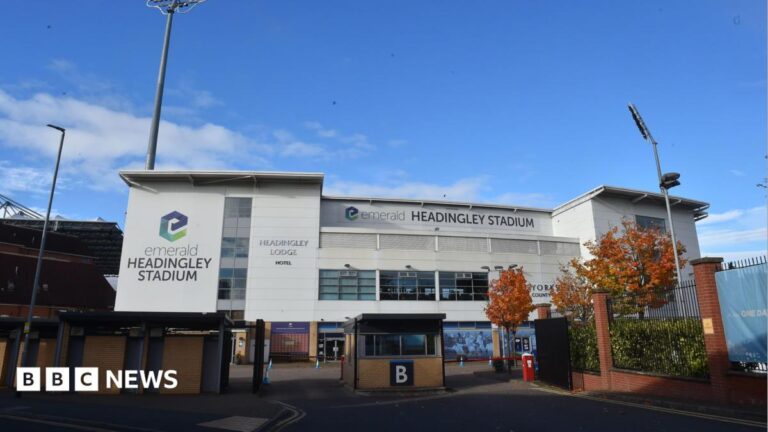 Headingley Stadium cancer centre plans approved The exterior of Headingley Stadium is shown with its large white panels, glass windows and tall floodlights rising above the building. A gated entrance and autumn‑coloured trees sit in the foreground, making the stadium look both modern and familiar as a major Leeds sports venue.