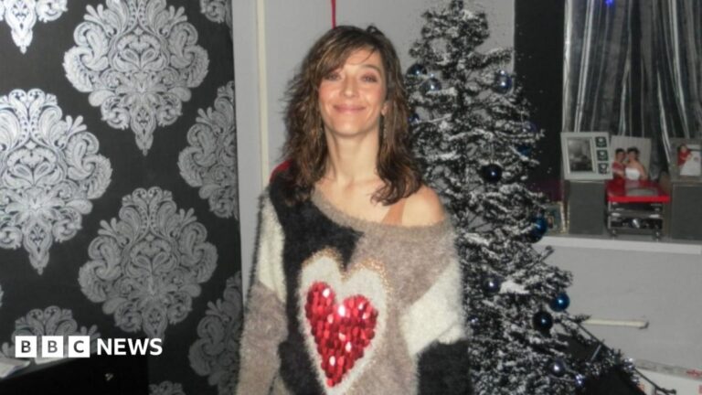 Is school to blame for mum's asbestos death? A woman with wavy shoulder length dark hair and an off the shoulder wooly jumper with a red love heart on the front stands smiling in front of a Christmas tree.