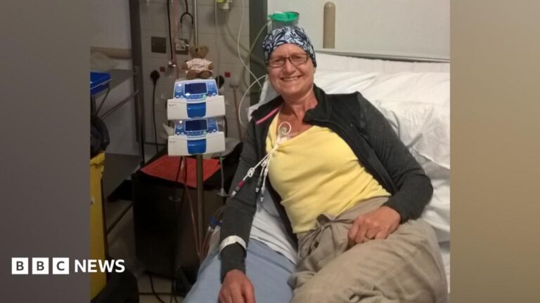 Maidstone woman’s early cancer symptoms blamed on menopause Sarah Chapman sits on a hospital bed wearing a patterned head covering and casual clothing. Medical equipment and infusion pumps are positioned beside the bed, with tubing attached to a line in Sarah Chapman's chest.