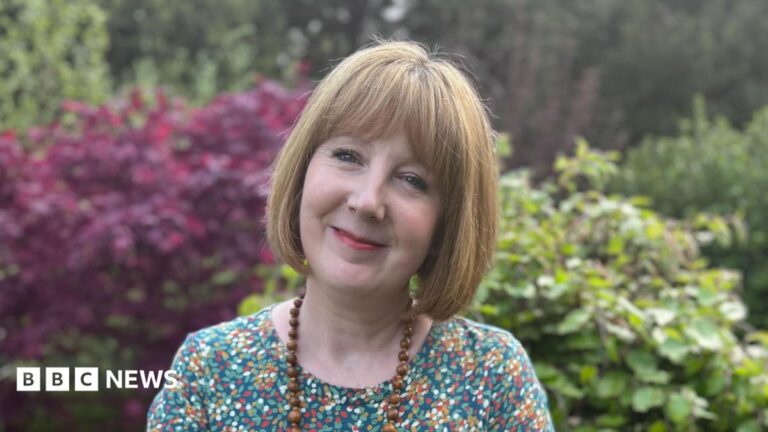 A woman with a short blonde bob and fringe smiles at the camera. She is wearing a multi-coloured T shirt and wooden bead necklace. Behind her are red and green bushes.