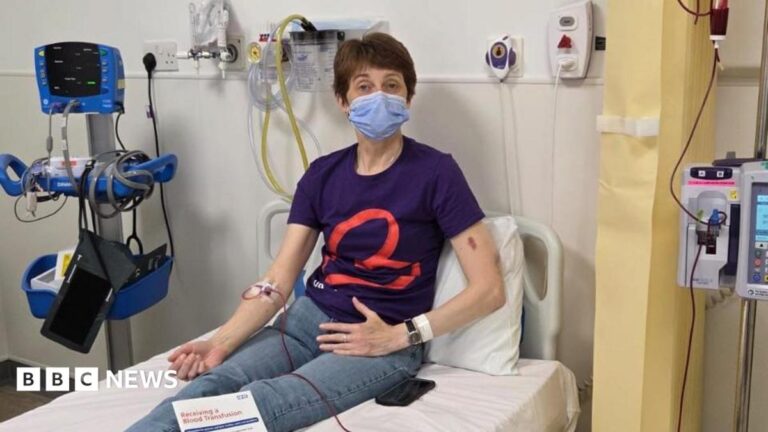 A woman with short dark hair and wearing a face mask sits on a hospital bed in blue T-shirt and jeans, hooked up to a red tube connected to a transfusion machine.
