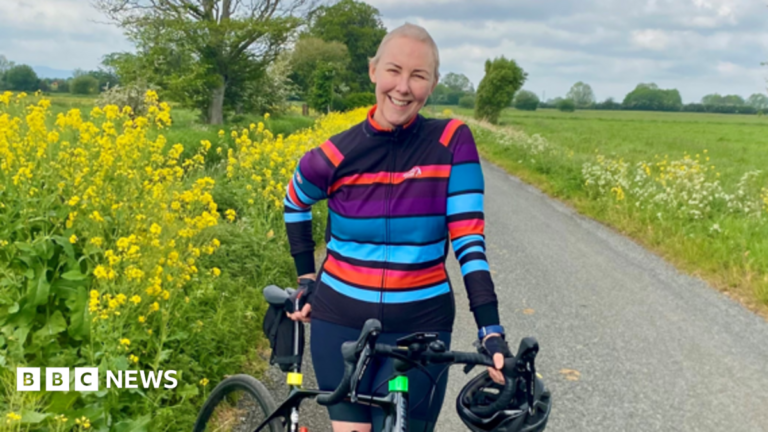 Somerset woman ‘saved’ by thyroid cancer trial rides for hospital Julie smiling to the camera on a country lane with green fields, trees and cow parsley in the background. She is wearing a striped blue and mauve cycling jacket and is leaning against her black bike with a helmet in her hand.