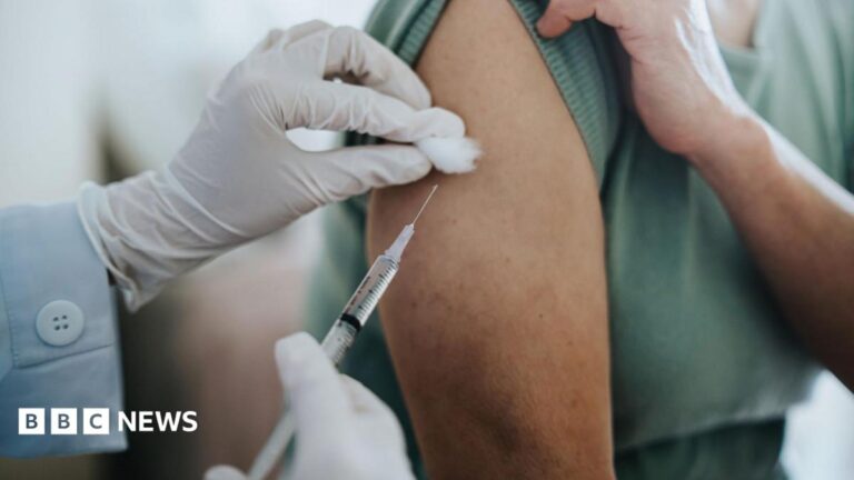 Close-up of a healthcare professional administering an injection into a person's upper arm, with white gloved hands holding a syringe and cotton swab during a vaccination appointment.