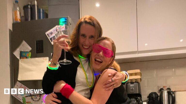 Two women embracing and laughing. They are hugging and wearing colourful accessories. One has a big glass of white wine in her hand.