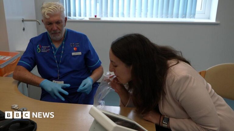 A woman with long dark hair and wearing a light pink jacket leans forward towards a desk.  She is blowing into a tube on a clear plastic bag which is attached to a small white machine with a built in monitor.  To her left is a nurse watching her progress. He is wearing royal blue scrubs and blue latex gloves and has white hair and a short white beard.