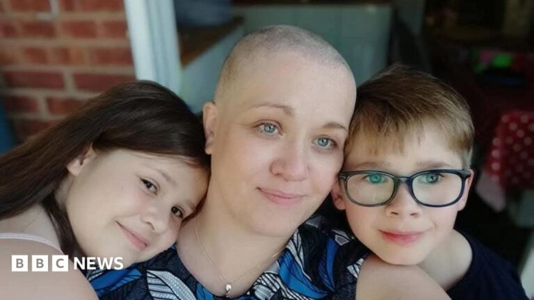 ‘A tumour the size of a football saved my life’ A woman who has lost her hair is being hugged by two children. They appear to be standing outside a house.