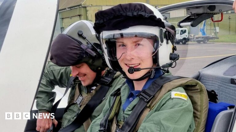Matthew Adams sitting in the cockpit of a plane, wearing a green jumpsuit and a white helmet with a protective guard across his eyes and communications microphone in front of his mouth. He smiles towards the camera whilst a man sitting next to him straps himself into the seat.