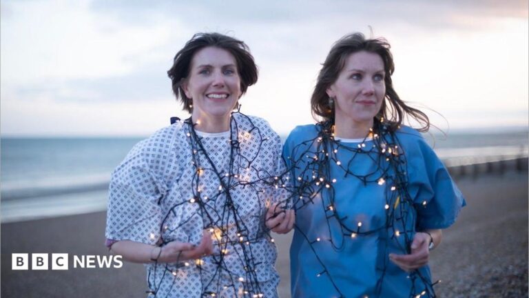 Hastings twin sisters turn life and illness into comedy play A woman with short hair and a patterned white shirt is standing next to a long-haired woman in a blue shirt. They are standing on a beach and tangled in string lights.