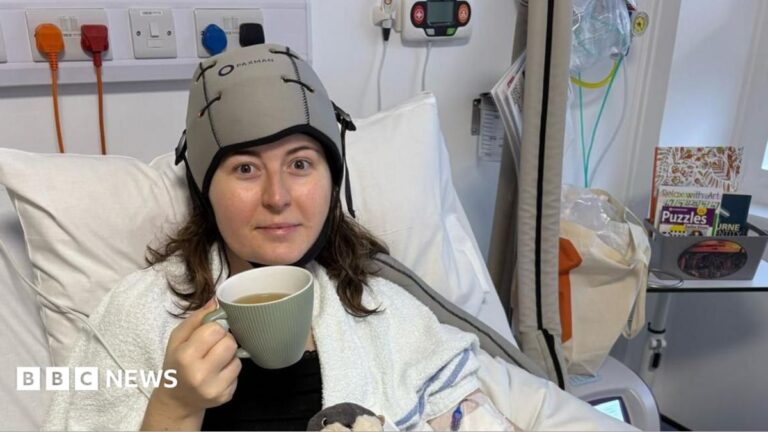 Fionnuala Quinlan-Bennett is seen in a hospital treatment area. She is sitting upright in a hospital bed. A cooling cap is fitted to her head and tubing is attached to medical equipment. A large mug, books and personal belongings are visible nearby. Medical monitoring equipment and hospital furnishings can be seen in the background.