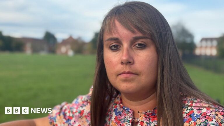 Becky Jones has shoulder-length brown hair with a fringe. She is standing in a park and is wearing a dress with multi-coloured flowers on it.