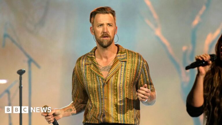 Charles Kelley of Lady A performing onstage during the 2024 People's Choice Country Awards at Grand Ole Opry House in Nashville, Tennessee. He is wearing a gold stripey shirt and holding a microphone.