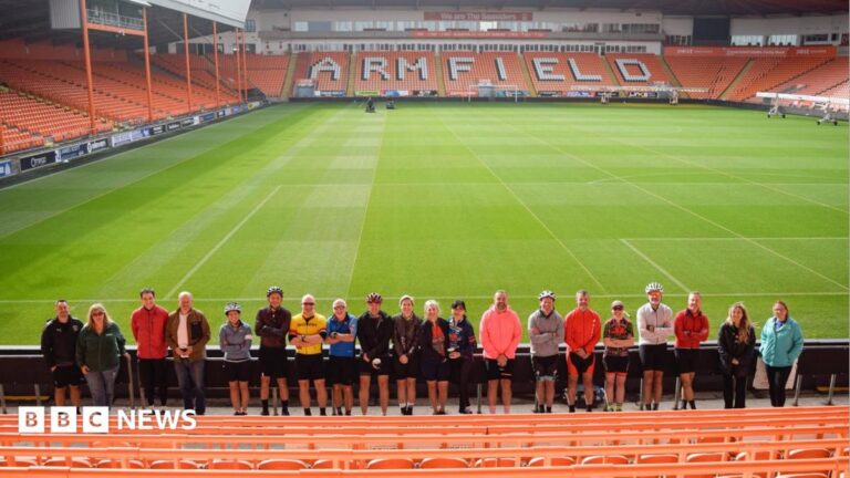 Lancashire cancer clinicians complete 140-mile charity bike ride A large group of people with cycling helmets smile as they stand in front of a football pitch. They are standing in a long row, almost all of them wearing black shorts and with an assortment of coloured tops. In front of them are rows of orange seats, behind them the green pitch. Beyond that are further rows of orange seating with the word ARMFIELD written in white capital letters across the seats.