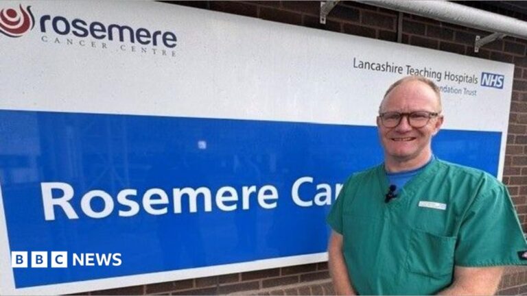 Lancashire cancer clinicians take on 140-mile charity bike ride Jeremy Ward wears green surgical scrubs while standing outside a hospital building. He has short grey hair and wears glasses.