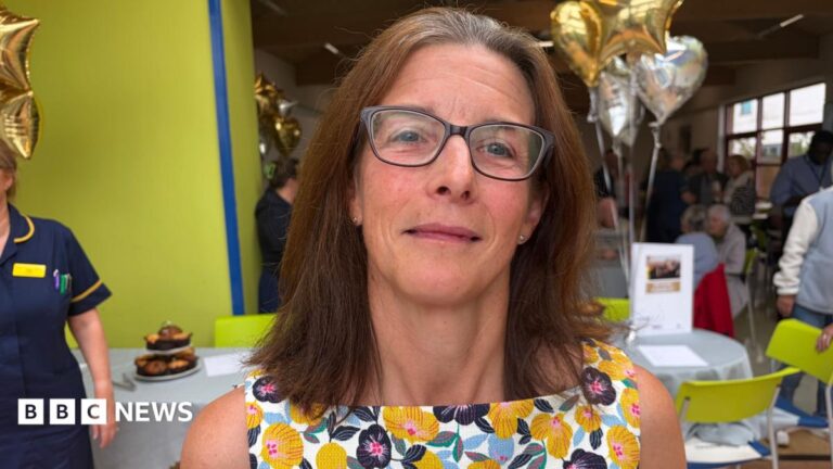 A woman with shoulder-length brown hair and grey roots is wearing black glasses and has a white top on with yellow, blue and black flowers on it. In the background are silver heart balloons and gold star balloons