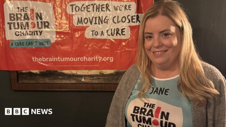 Somerset mum mistook brain tumour symptoms for perimenopause Jane is wearing a Brain Tumour Charity T-shirt and is stood in front of a similar banner on a wall behind her. She has long blonde hair and is smiling at the camera.