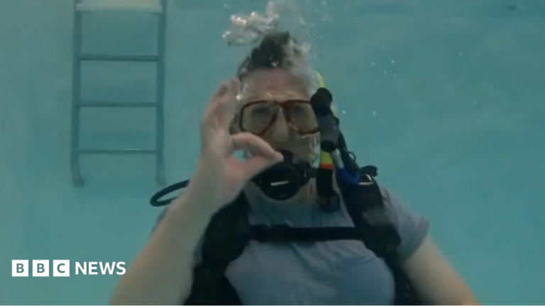 Luke Rogers is seen submerged in a pool, wearing a scuba mask and diving equipment. He is staring at the camera and making an expression with his fingers and hand to indicate things are going well