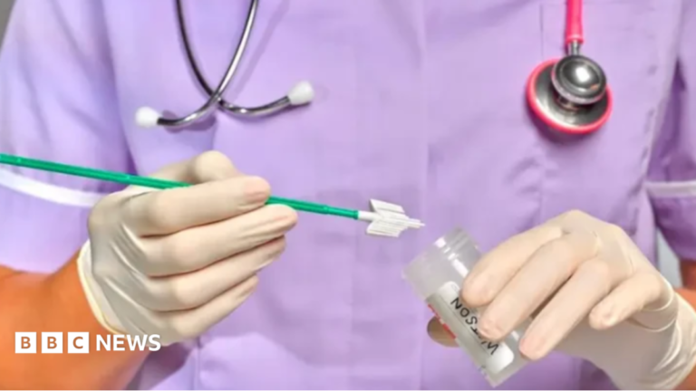 A woman in white rubber gloves holds a test in one hand and a pot to deposit it in the other.  A stethoscope hangs round her neck and she wears a purple uniform.