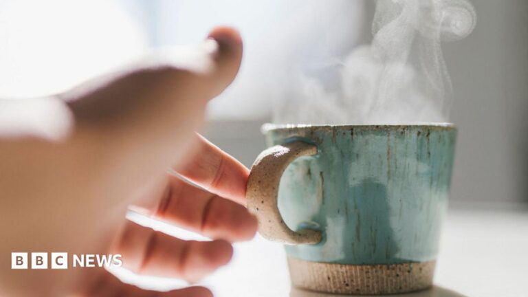 A woman reaches her hand out to pick up a steaming hot mug which is shiny and green-coloured