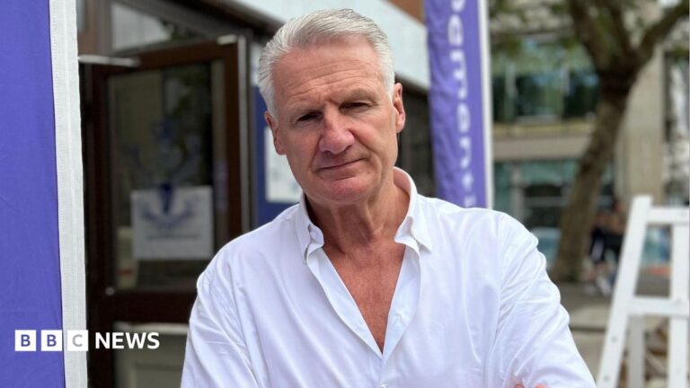 Tom Binet pictured in a white shirt. He has short cropped white hair. He has a serious expression. He is outside and there are some buildings out of focus behind him.